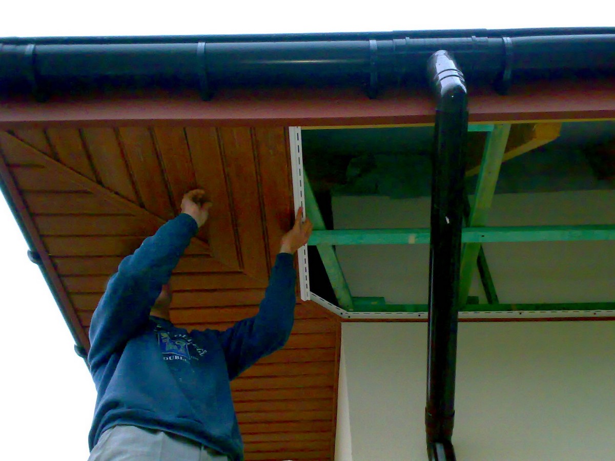 Pracownik montuje drewnianą podbitkę dachową, widoczne fragmenty konstrukcji dachu i rynna spustowa.