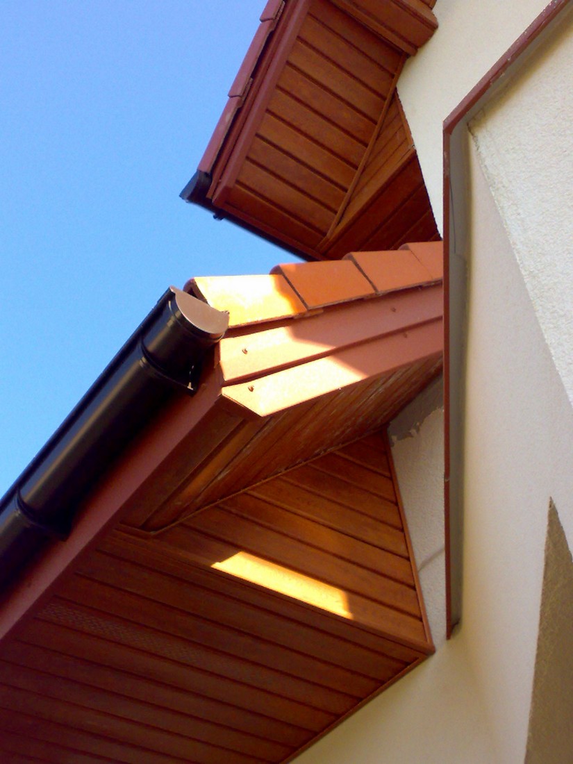 Wykończona podbitka dachowa z brązowego drewna, widok z dołu na róg budynku z zamontowaną rynną, jasne słońce podkreśla strukturę drewna.