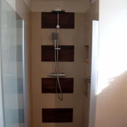 Wykończona kabina prysznicowa z deszczownicą i baterią termostatyczną na ścianie z beżowych płytek z ciemnobrązowymi wstawkami, widoczny odpływ liniowy i półka w ścianie.