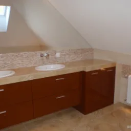 Nowoczesna łazienka na poddaszu ze skośnym sufitem, wyposażona w dwie białe umywalki nablatowe, brązowe szafki z szufladami, mozaikę z beżowych płytek nad blatem, grzejnik żeberkowy oraz okno...