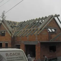 Wznoszenie domu z cegły, widoczna konstrukcja drewniana dachu w trakcie budowy, z przodu fragment samochodu z logo firmy dekarsko-ciesielskiej.