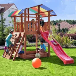 Drewniany plac zabaw z kolorowym daszkiem, zjeżdżalnią i drabinką, otoczony zielonym trawnikiem, na którym bawią się dzieci. Widoczne domy w tle.