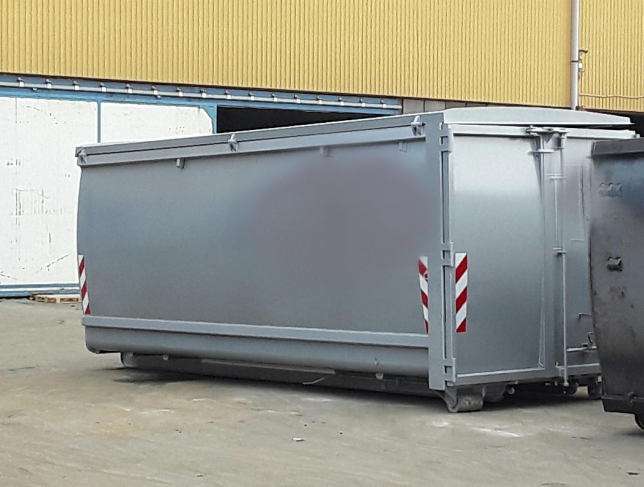 Szary, metalowy kontener z czerwono-białymi pasami ostrzegawczymi, stojący na betonowym podłożu przed żółtym budynkiem z przesuwanymi drzwiami.