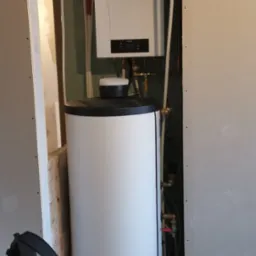 Biały bojler i piec gazowy z niebieskim zbiornikiem wyrównawczym zamontowane w przestrzeni między ścianami z płyt gipsowo-kartonowych, widoczne rury instalacji i czarny plecak na podłodze.