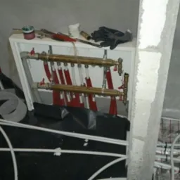 Rozdzielacz ogrzewania podłogowego w trakcie montażu, widoczne rury grzewcze na podłodze z siatką, narzędzia na półce, surowe ściany w tle.