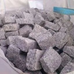 Szare kostki granitowe w worku typu big bag, widoczne niebieskie uchwyty. Kostka o nieregularnych kształtach wypełnia worek.