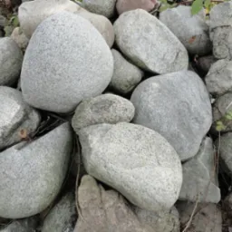 Stos szarych, gładkich kamieni różnej wielkości, tworzących naturalną kompozycję z elementami zieleni w tle. Kamienie o zróżnicowanej fakturze i kształtach.
