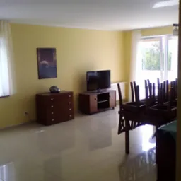 Salon z żółtymi ścianami i błyszczącą, kremową podłogą z płytek w Poznaniu. Widoczne meble: komoda, szafka pod telewizor, stół z krzesłami ustawionymi do góry nogami. Okno z białymi zasłonami...