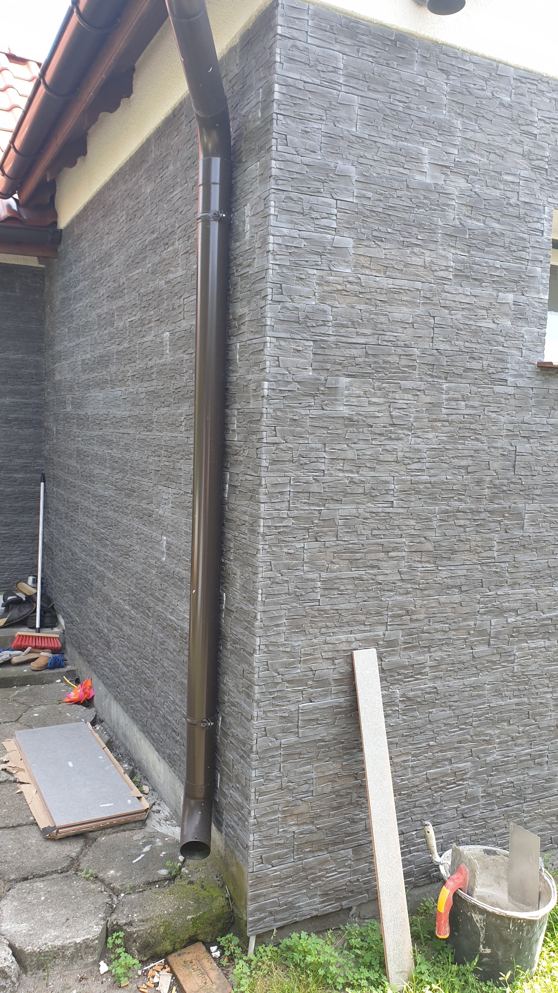 Szara elewacja budynku wykończona płytkami imitującymi kamień, z brązową rynną i narzędziami glazurniczymi w pobliżu, wskazującymi na trwający montaż.