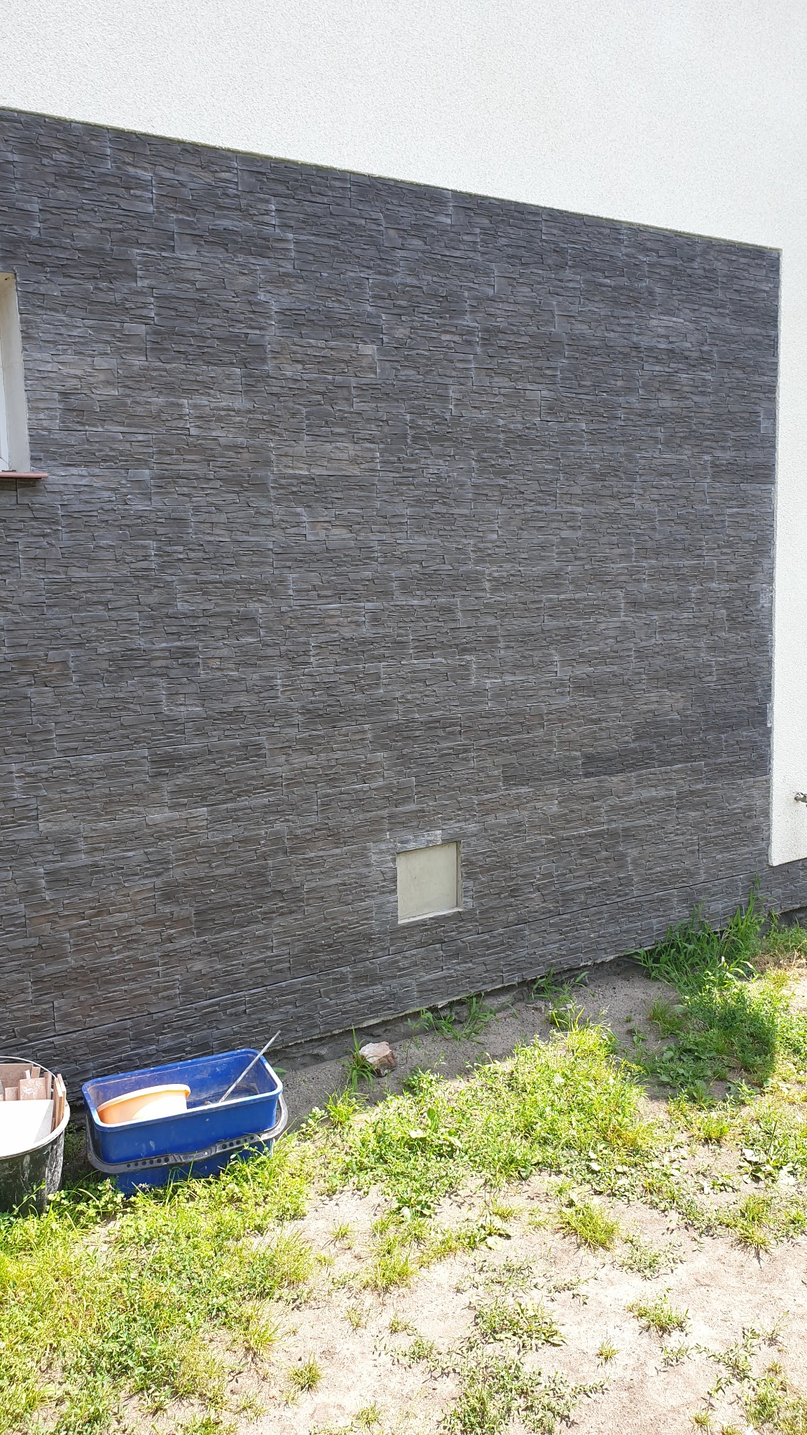 Elewacja budynku wykończona szarą okładziną kamienną, widoczne narzędzia i pojemniki malarskie na trawie u podstawy ściany.