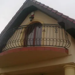 Kuty balkon z dekoracyjnymi elementami na tle beżowej ściany domu z brązowym dachem.