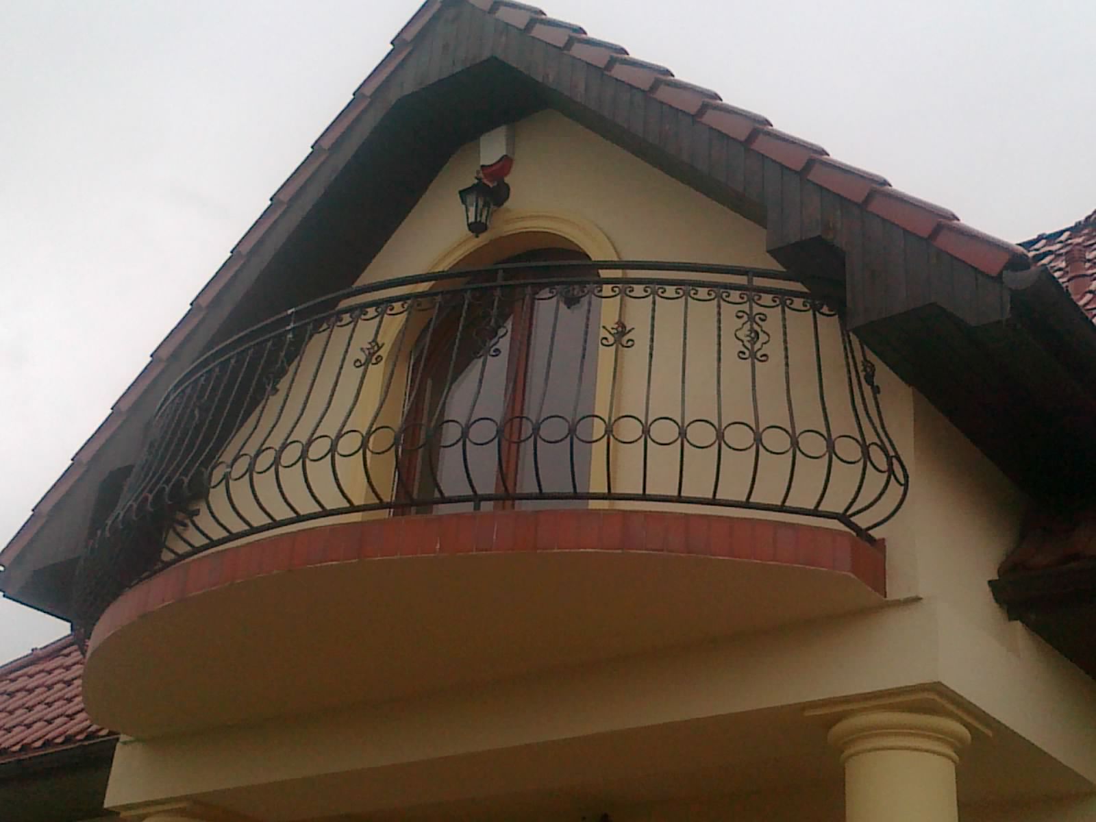 Kuty balkon z dekoracyjnymi elementami na tle beżowej ściany domu z brązowym dachem.