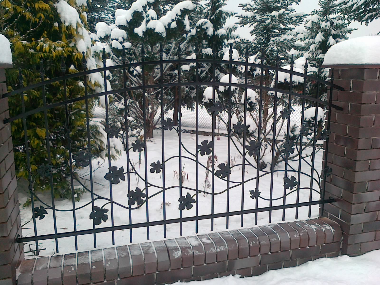 Kute ogrodzenie z motywem liści winogron, osadzone na ceglanym murku, zimowy krajobraz w tle, drzewa pokryte śniegiem.