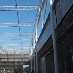 Wnętrze hali w budowie z widoczną stalową konstrukcją szkieletową i fragmentem ściany z płyt warstwowych. Na dole widoczni pracownicy w odblaskowych kamizelkach.