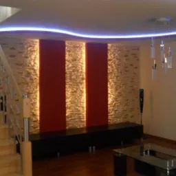Salon z elementami dekoracyjnymi: podświetlana listwa LED na suficie, ściana z kamienia dekoracyjnego z dwoma pionowymi pasami w kolorze czerwonym, drewniane schody z metalową balustradą, szklany...