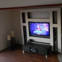 Telewizor wbudowany w ścianę z szafką RTV i głośnikami, aranżacja wnętrza z drewnianą podłogą i dekoracyjnymi wazonami.