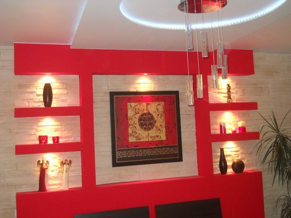 Czerwona ściana z półkami i oświetleniem punktowym, dekorowana obrazem w ramie, wazonami i figurkami, na tle ściany z imitacji kamienia, z nowoczesnym żyrandolem.