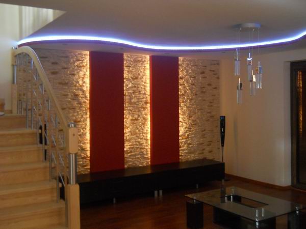 Salon z elementami dekoracyjnymi: podświetlana listwa LED na suficie, ściana z kamienia dekoracyjnego z dwoma pionowymi pasami w kolorze czerwonym, drewniane schody z metalową balustradą, szklany...