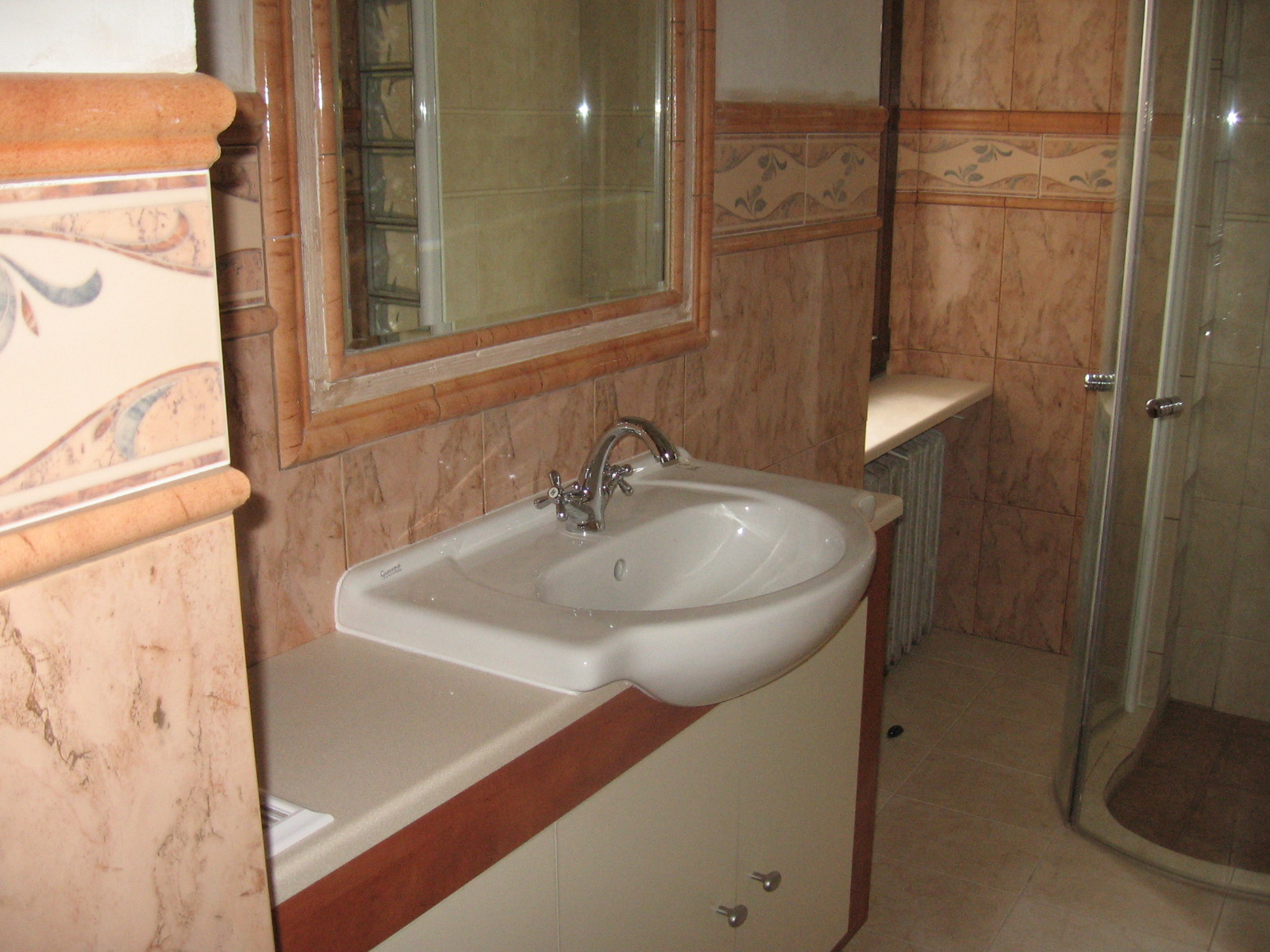 Wykończona łazienka z umywalką nablatową, lustrem w ozdobnej ramie i kabiną prysznicową, wyłożona płytkami w odcieniach beżu i brązu z dekoracyjnymi bordiurami.