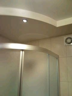 Łazienka po remoncie: kabina prysznicowa z matowego szkła, jasne płytki na ścianach, sufit podwieszany z oświetleniem punktowym i wentylator w rogu.