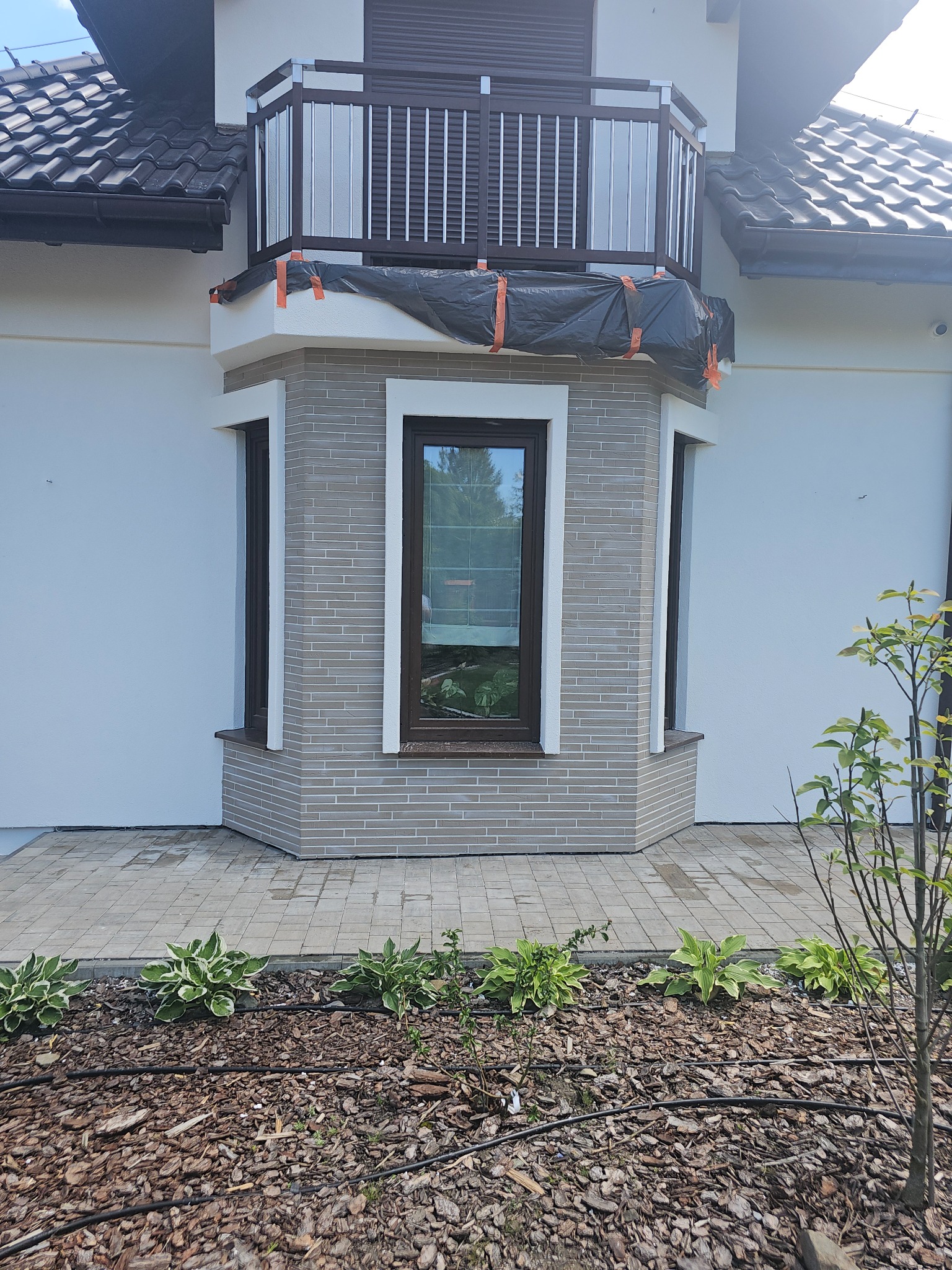 Wykończona elewacja domu z oknem wykuszowym obłożonym szarą cegłą. Balkon z brązowymi żaluzjami zabezpieczony folią. Widoczne rośliny wzdłuż ścieżki.