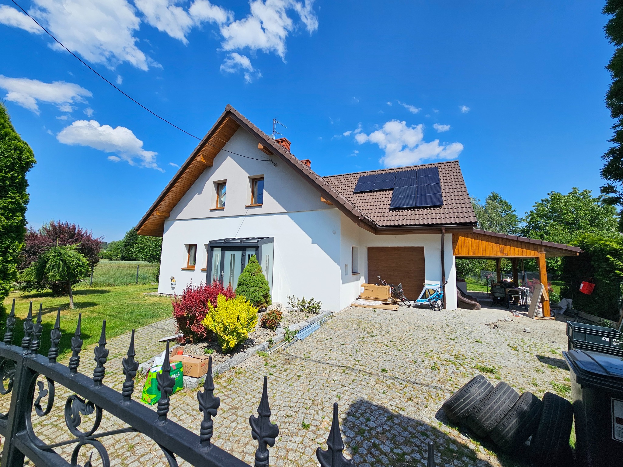 Dom jednorodzinny z białą elewacją, brązowym dachem z panelami słonecznymi i kolorowym ogrodem, widziany zza ozdobnego, metalowego ogrodzenia.