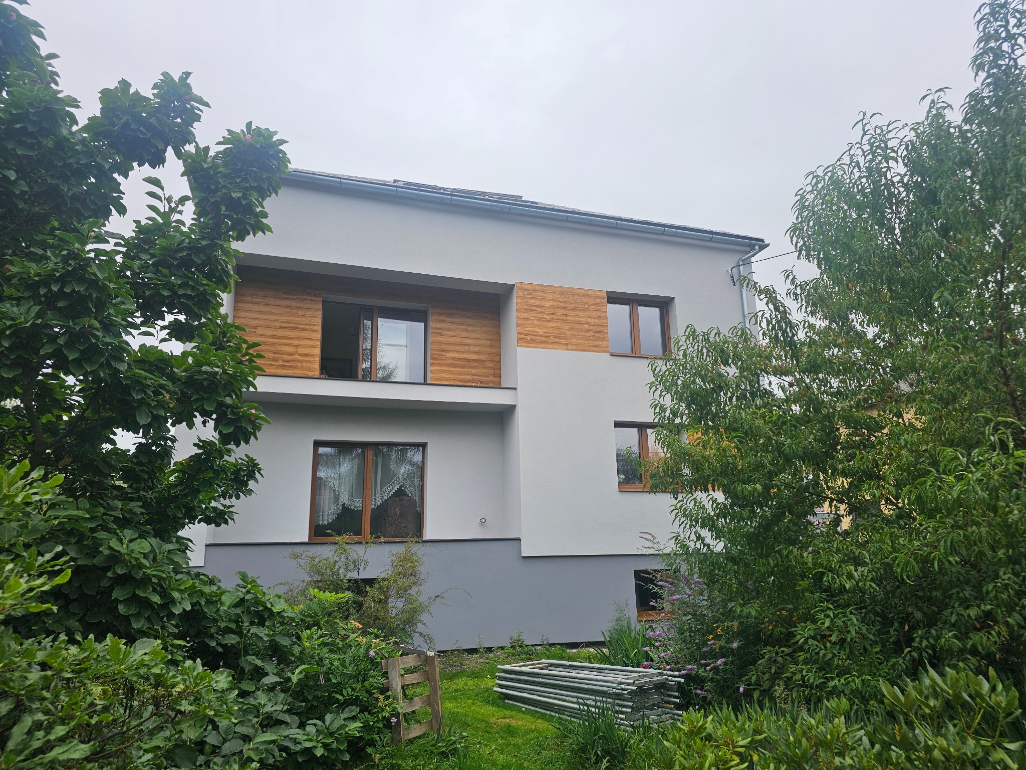 Dwukondygnacyjny dom z szarą elewacją i drewnianymi panelami na balkonie, otoczony zielenią. Na trawniku leżą złożone rusztowania.