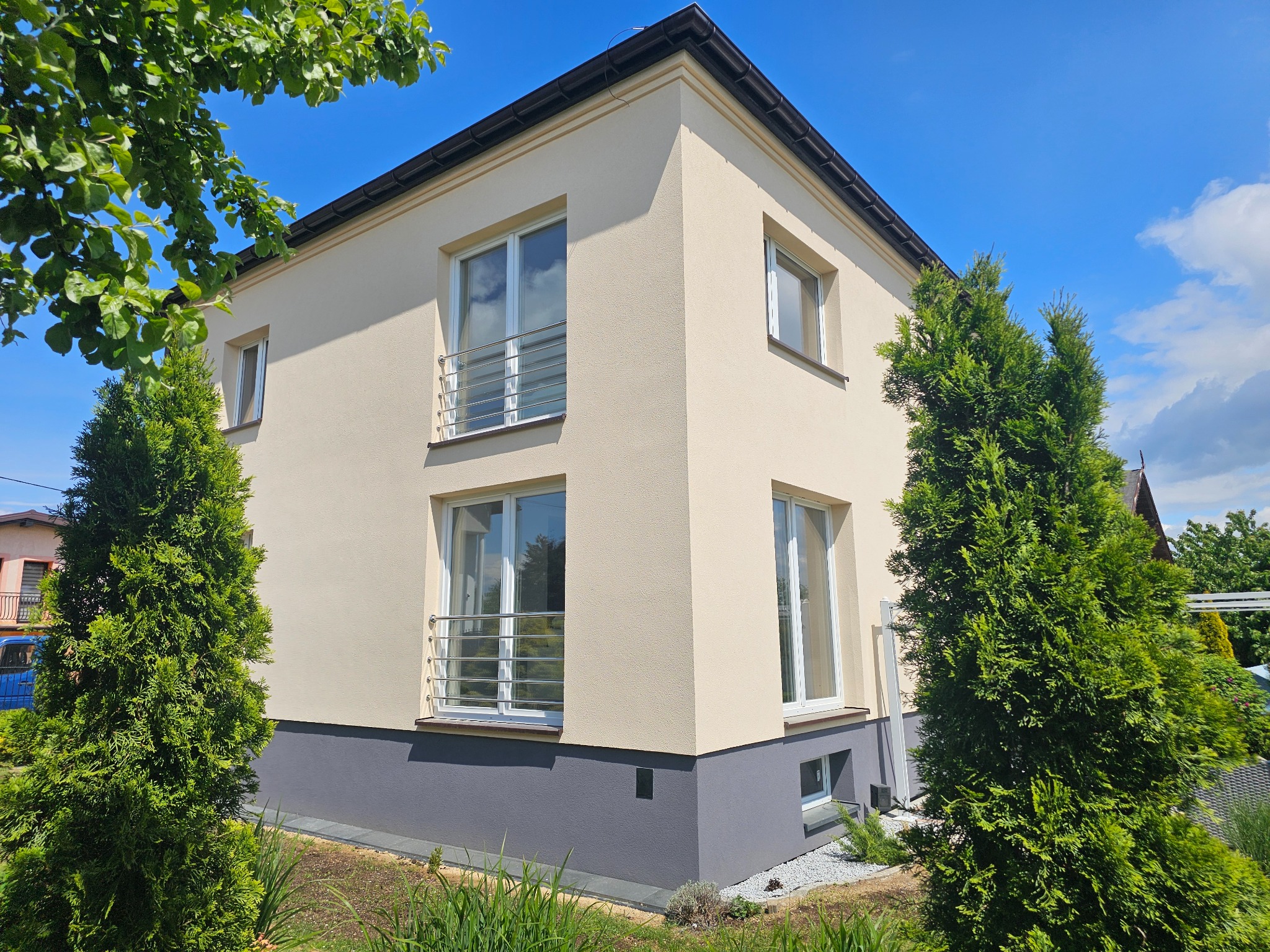 Dwukondygnacyjny dom z beżową elewacją i szarym cokołem w Leśnej, otoczony zielenią. Widoczne okna z balustradami i fragment nieba.