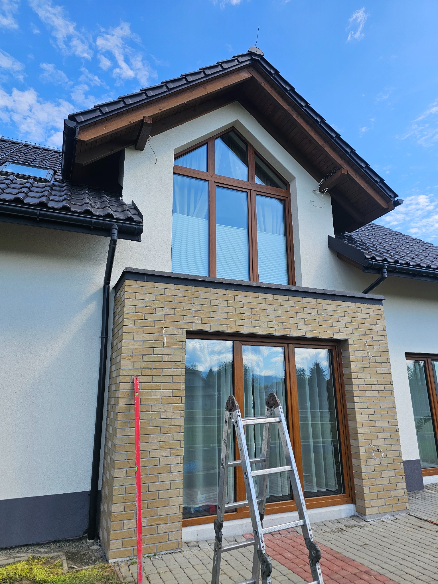 Dwukondygnacyjny dom z jasną elewacją i ceglaną wstawką, widoczna drabina i poziomica, sugerujące prace wykończeniowe. Ujęcie z dołu, błękitne niebo w tle.