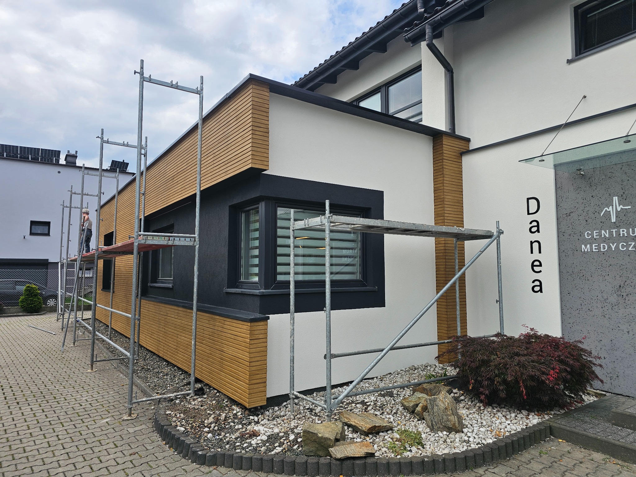 Nowoczesna elewacja budynku z elementami drewnianymi i białym tynkiem, widoczne rusztowania, sugerujące prace wykończeniowe.