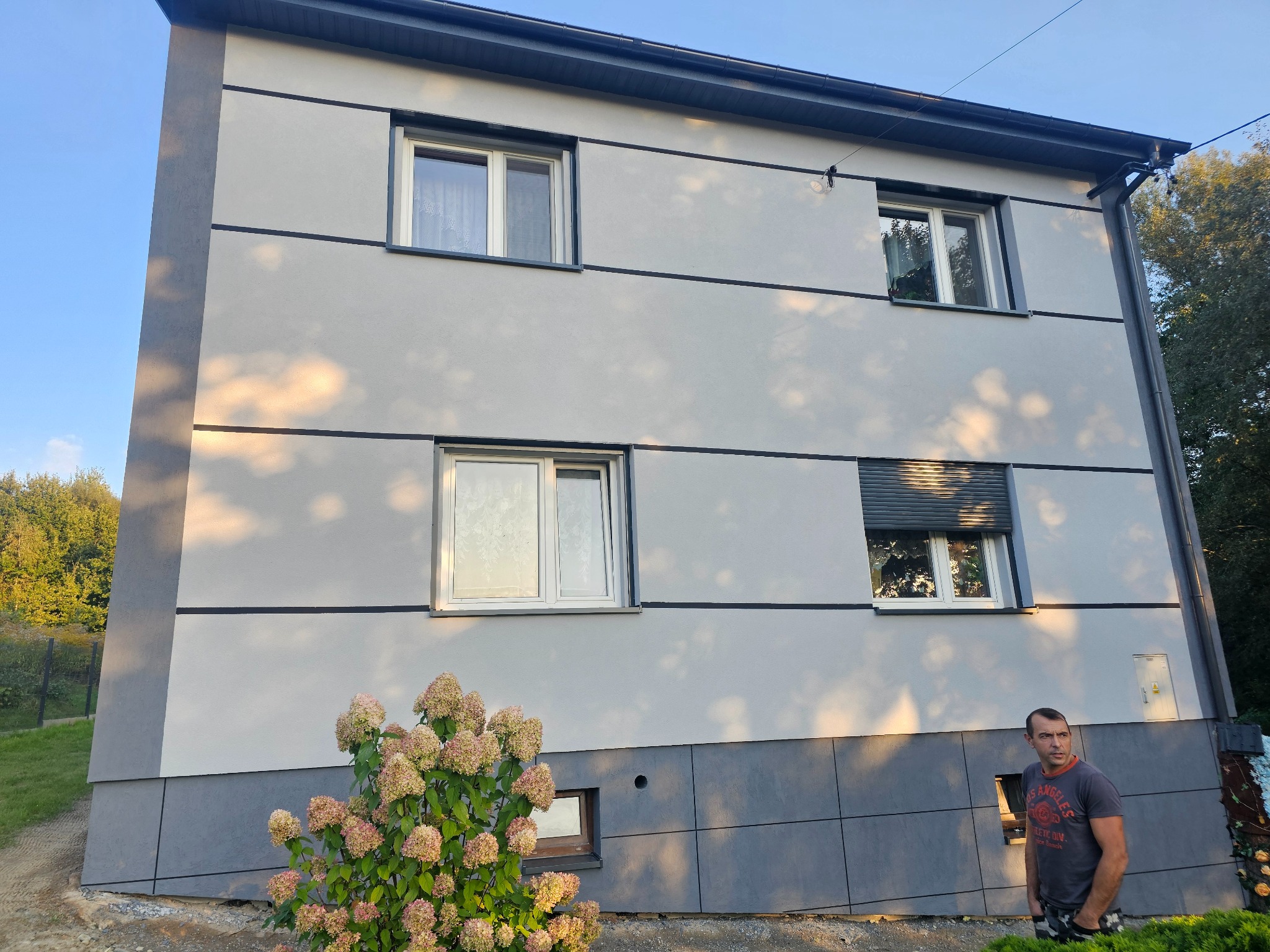 Dwukondygnacyjny dom z szarą elewacją, ciemnymi akcentami i widoczną roślinnością. Mężczyzna stoi obok budynku. Widoczne okna z roletami i bez.