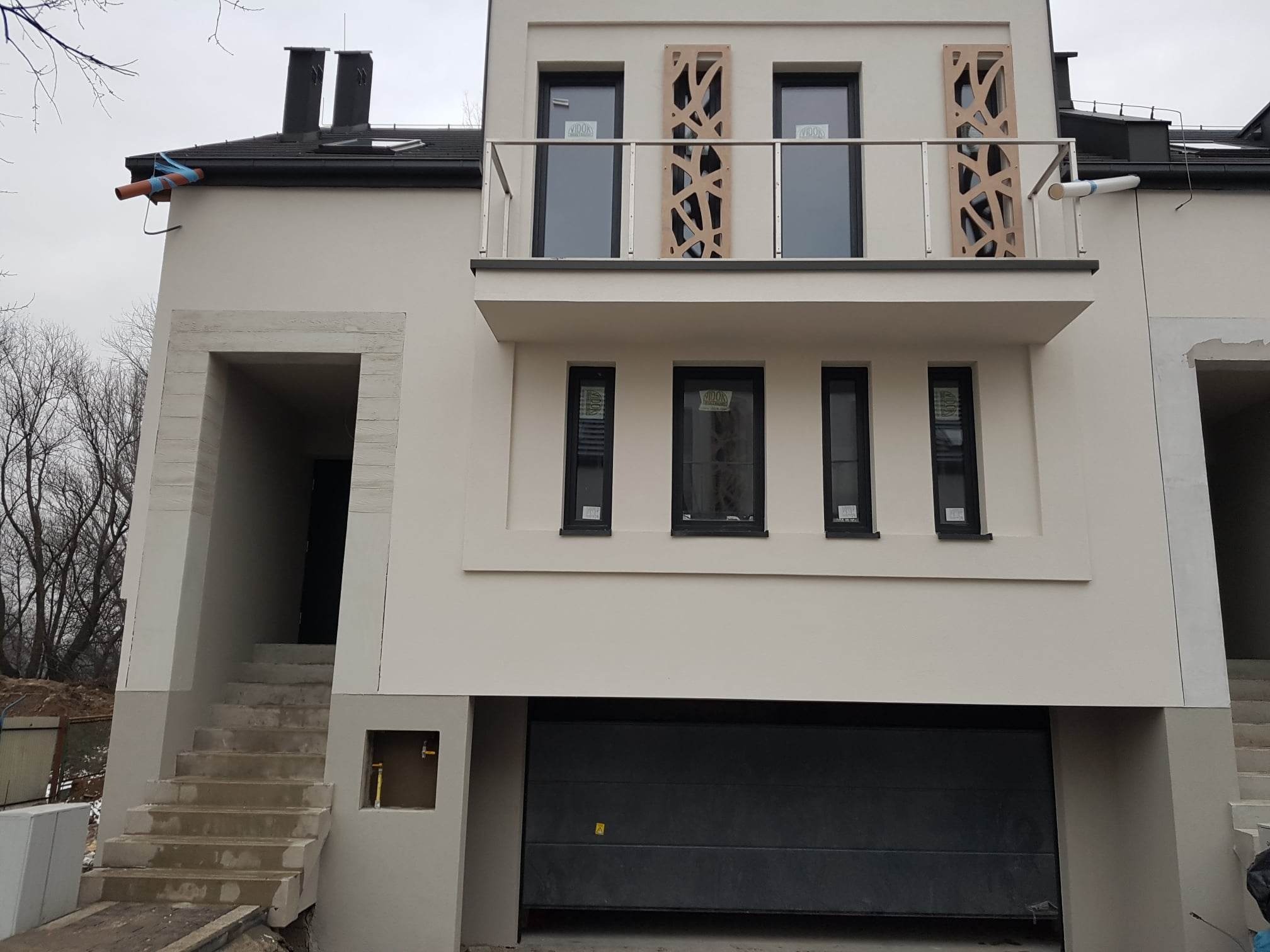 Nowoczesny dom z beżową elewacją, balkonem z ażurowymi panelami i garażem. Widoczne schody wejściowe i elementy wykończeniowe.