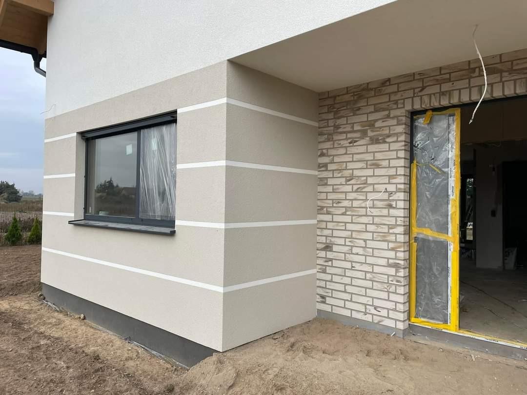 Wykończona elewacja domu z dekoracyjnym tynkiem w odcieniach beżu i białymi pasami. Fragment ściany wykończony cegłą. Okno zabezpieczone folią.