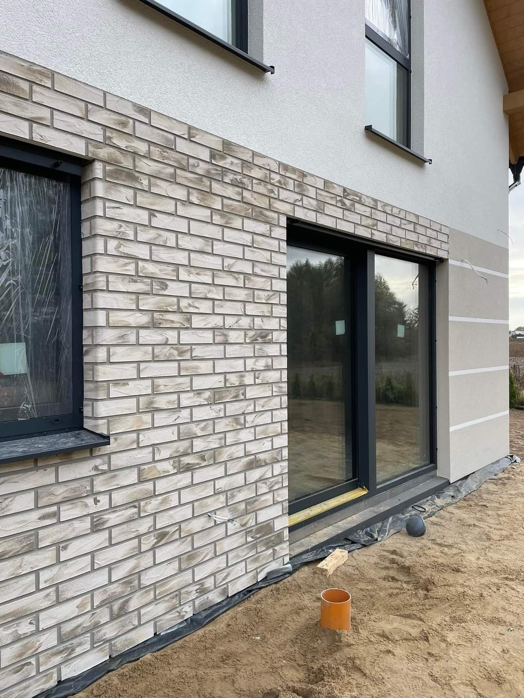 Elewacja domu jednorodzinnego z klinkieru w odcieniach bieli i szarości, widok z dołu, okna z czarnymi ramami, teren wokół domu w trakcie prac wykończeniowych.