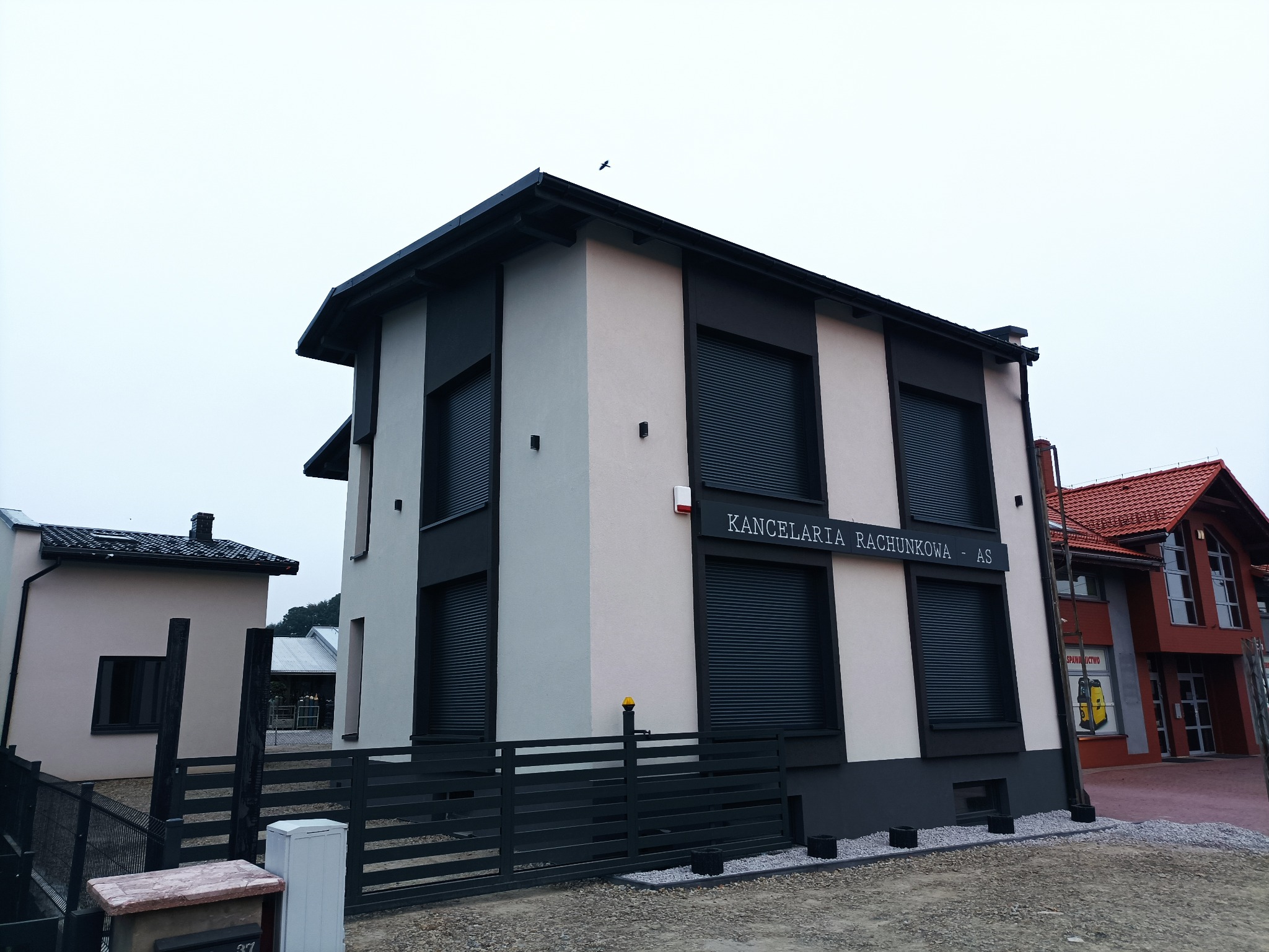 Dwukondygnacyjny budynek biurowy z elewacją w odcieniach szarości i czerni, z roletami zewnętrznymi i tablicą 'Kancelaria Rachunkowa - AS'.