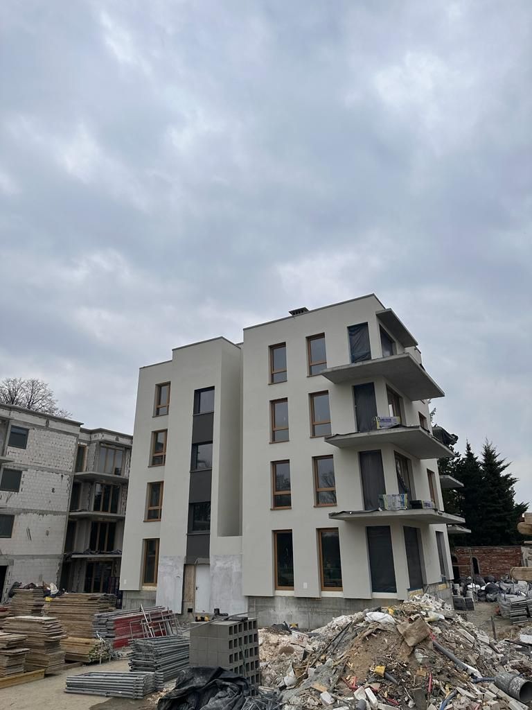Nowoczesny budynek wielorodzinny w trakcie prac elewacyjnych. Widoczne rusztowania, materiały budowlane i częściowo zakryte okna. Szara, pochmurna pogoda.