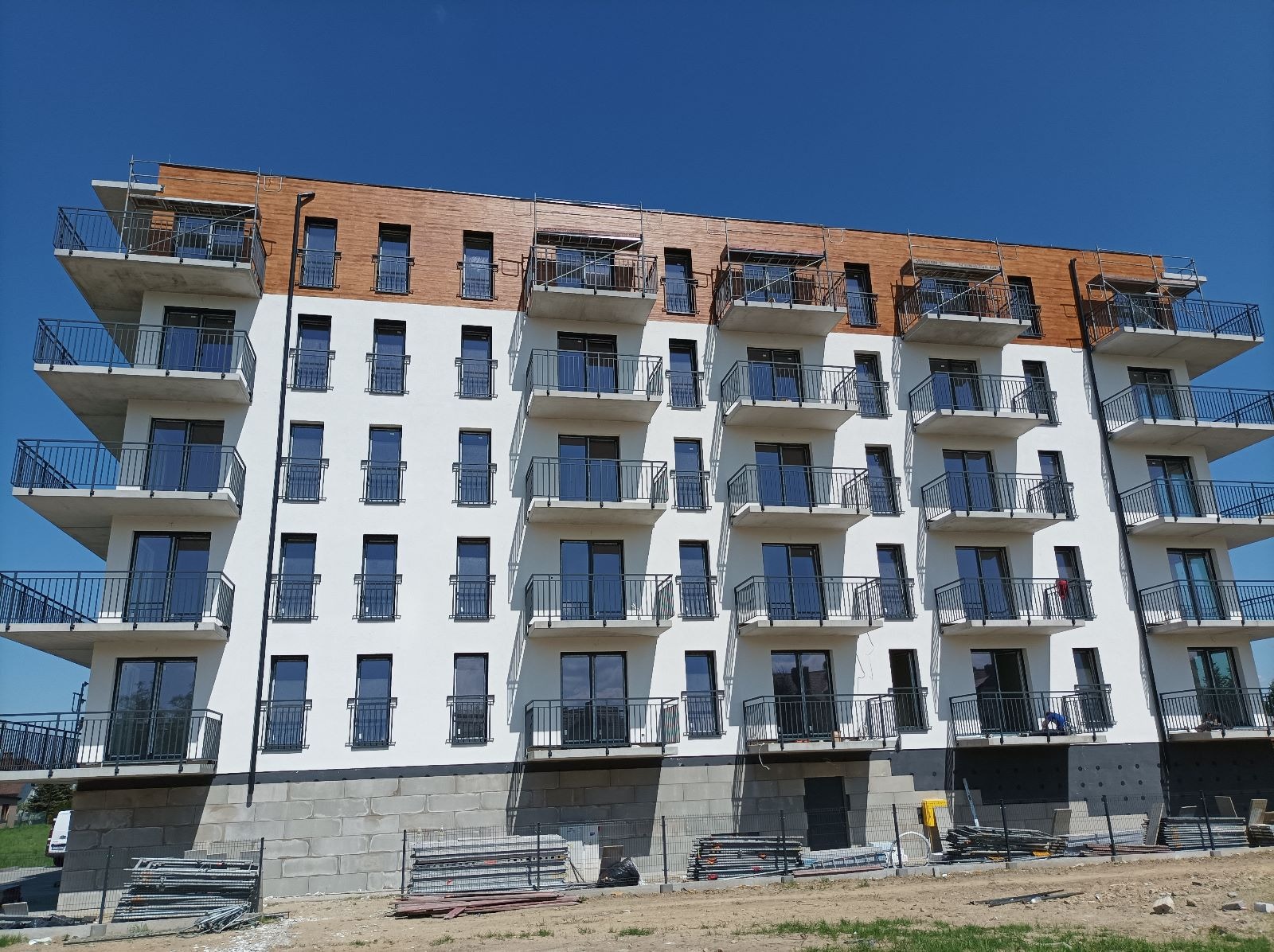 Nowoczesny budynek wielorodzinny z białymi ścianami i drewnianą górną częścią elewacji. Widoczne balkony z czarnymi balustradami i szare okna. Częściowo widoczne rusztowania.