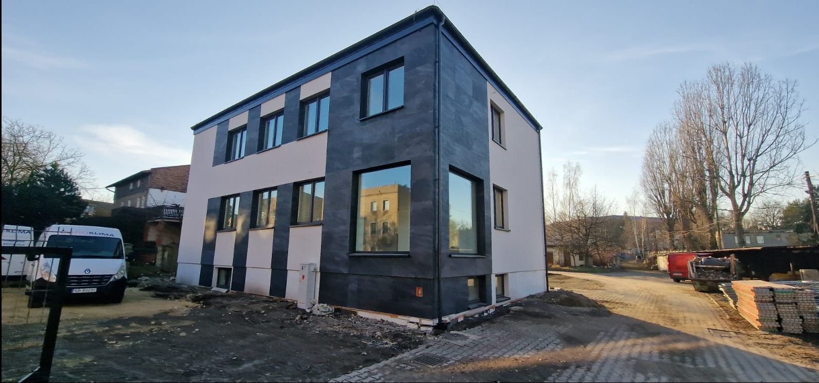 Dwukondygnacyjny budynek z nowoczesną elewacją w pasy, z przewagą szarości i bieli, widoczny z perspektywy ulicy w słoneczny dzień. W oknach odbija się sąsiedni budynek.