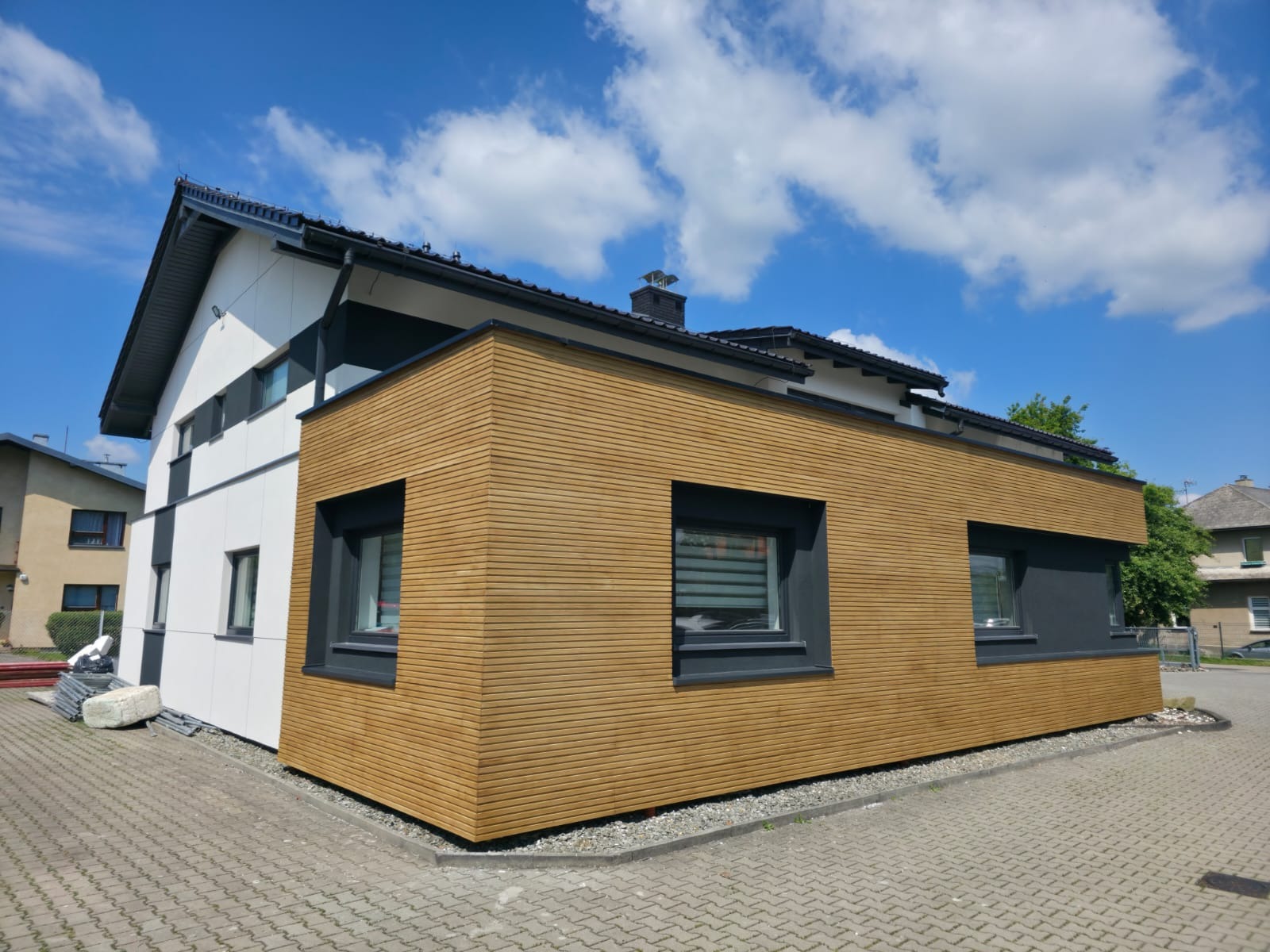 Nowoczesny dom z elewacją z drewnianych paneli i białym tynkiem, widok z zewnątrz w słoneczny dzień. Kontrastowe, ciemne obramowania okien.