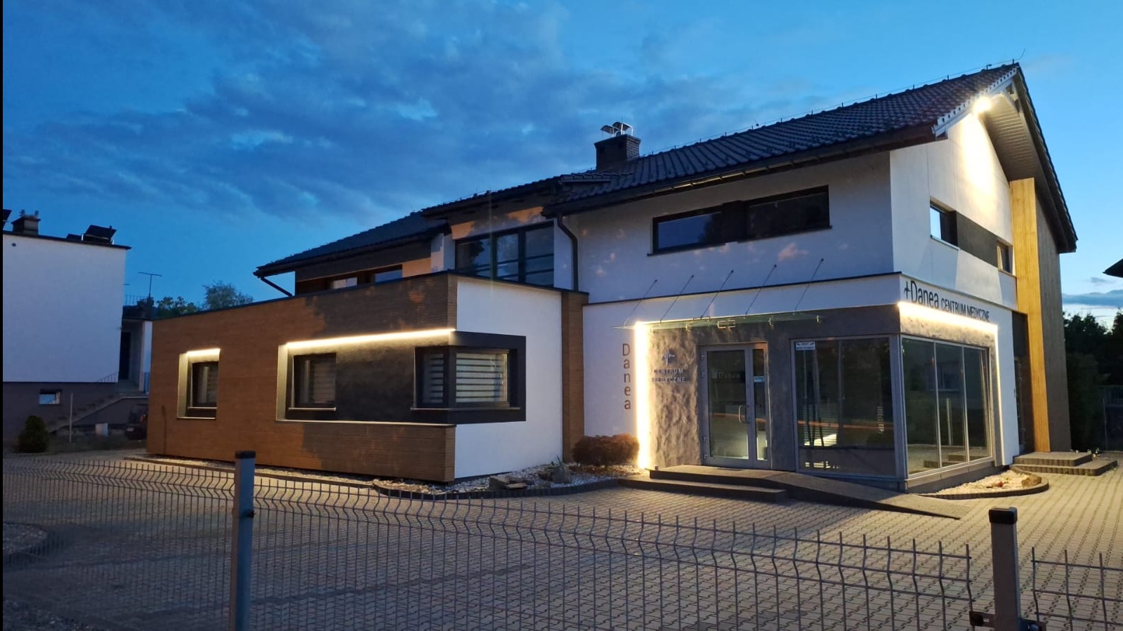 Nowoczesna elewacja budynku z oświetleniem LED, widoczna z zewnątrz. Jasna kolorystyka z elementami drewna i szarości. Ujęcie z poziomu gruntu, wieczorne oświetlenie.