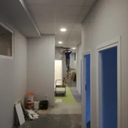 Pracownik na drabinie montuje sufit podwieszany w długim korytarzu biurowym z pomalowanymi na szaro ścianami i niebieskimi drzwiami, widoczne materiały budowlane i puste pojemniki po farbach.