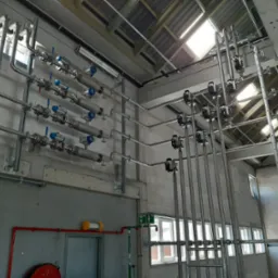 Instalacja rurociągów ze stali nierdzewnej z zaworami i osprzętem, zamontowana na ścianie z bloków betonowych w hali przemysłowej z widoczną konstrukcją dachu.