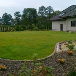 Rozległy trawnik przydomowy z równo przystrzyżoną trawą, obrzeżem z cegły i rabatami wysypanymi korą oraz ozdobnymi roślinami na pierwszym planie, w tle dom z szarą elewacją i drzewa.