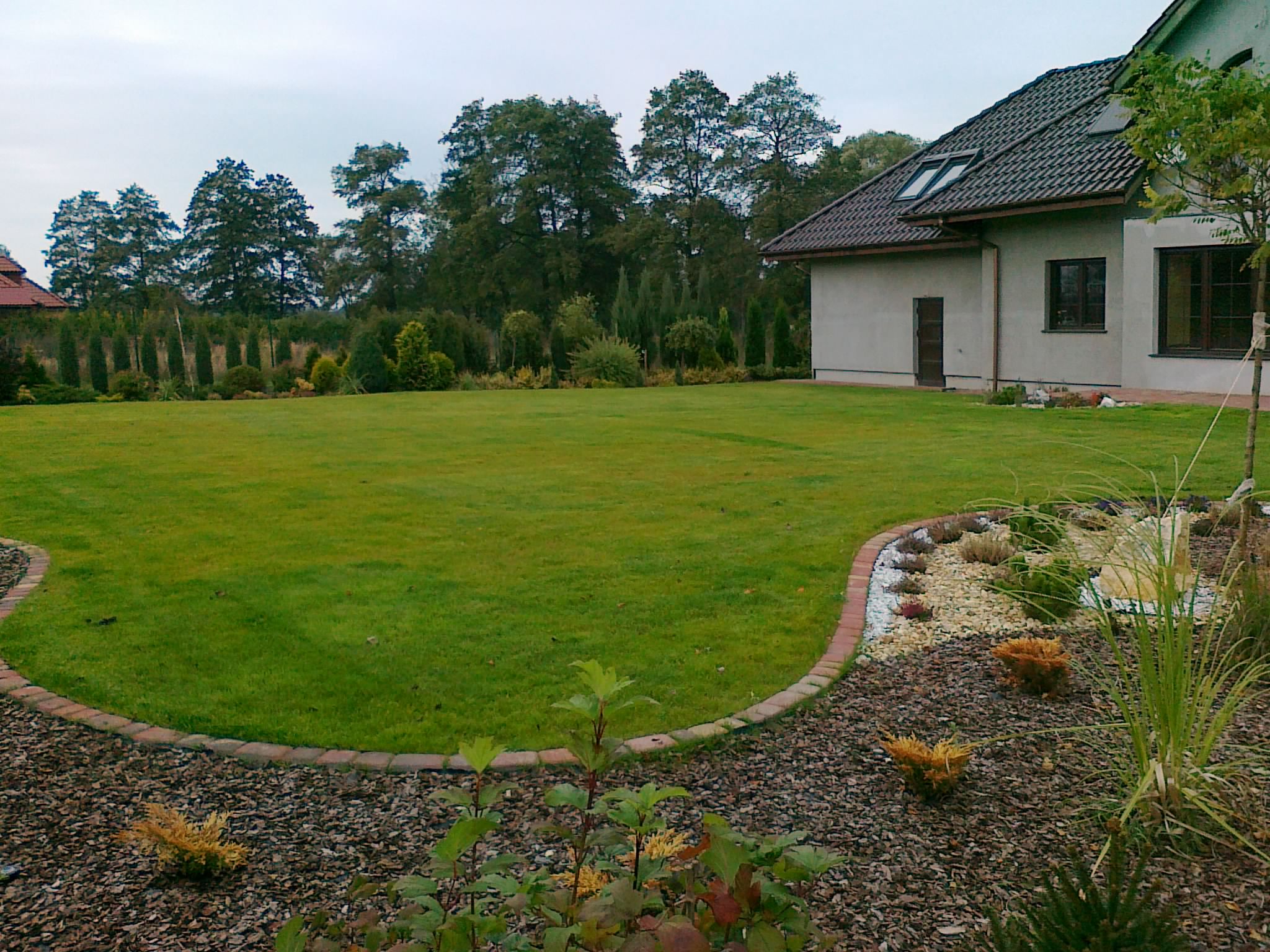 Rozległy trawnik przydomowy z równo przystrzyżoną trawą, obrzeżem z cegły i rabatami wysypanymi korą oraz ozdobnymi roślinami na pierwszym planie, w tle dom z szarą elewacją i drzewa.