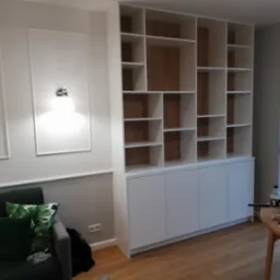 Biała zabudowa meblowa z szafkami dolnymi i otwartymi półkami w salonie, widoczny fragment szarej sofy z zielonymi poduszkami i oknem w tle.