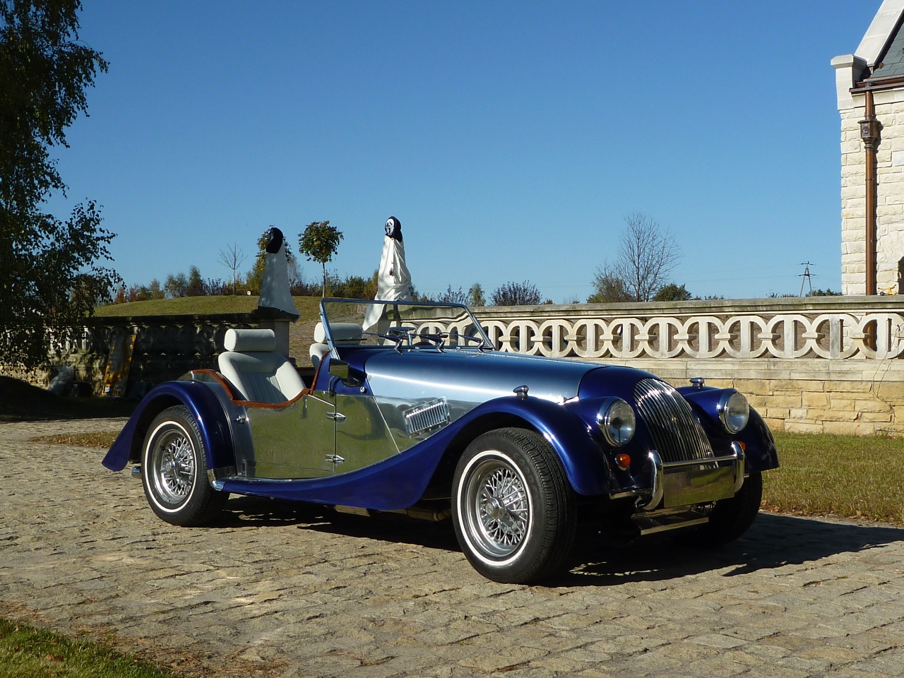 Elegancki, dwuosobowy, niebiesko-srebrny roadster z chromowanymi detalami, zaparkowany na brukowanej drodze przed kamiennym murem z ozdobnymi elementami, w tle drzewa i błękitne niebo.