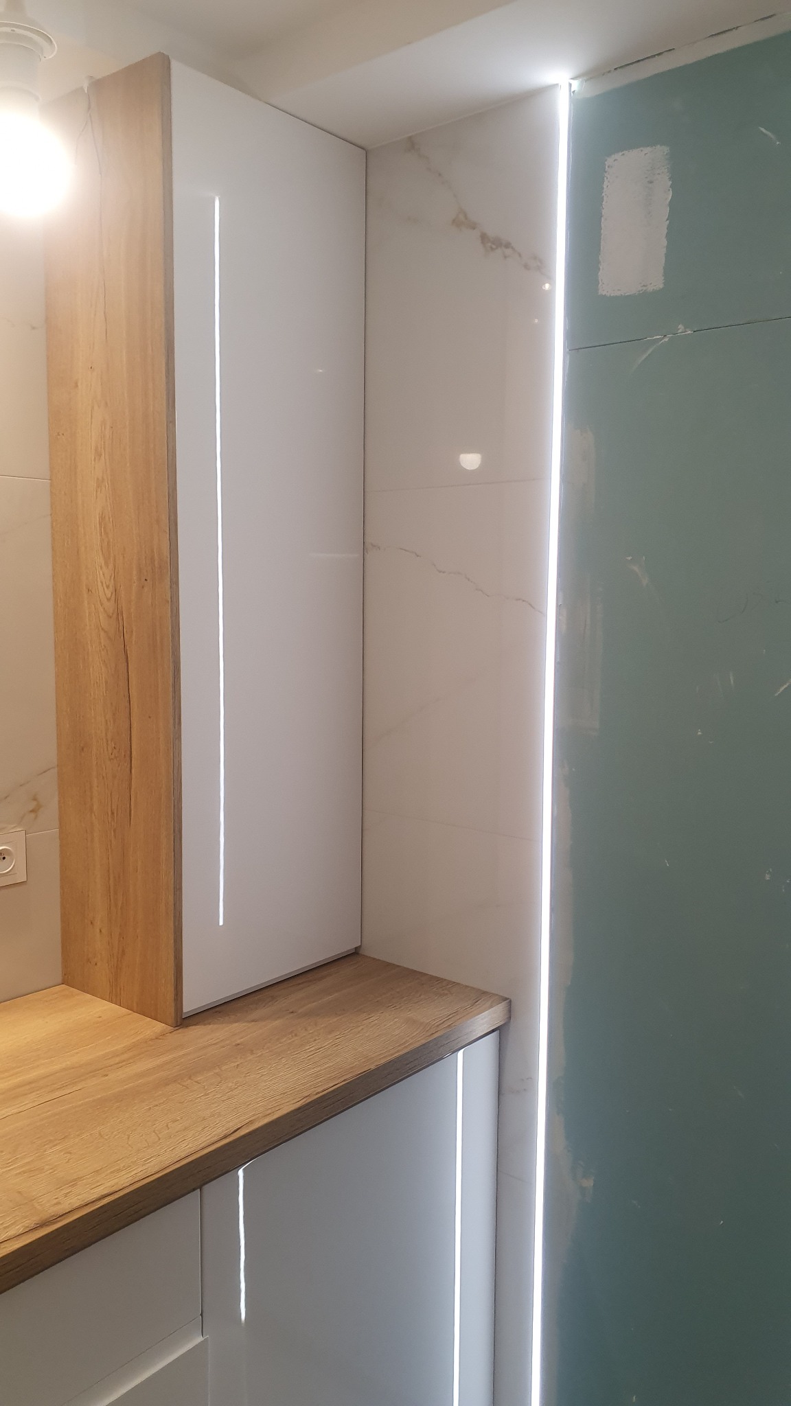 Fragment zabudowy kuchennej z białymi frontami i drewnianym blatem, oświetlenie LED w pionowych szczelinach, marmurowe płytki na ścianie, widoczny fragment surowej ściany.