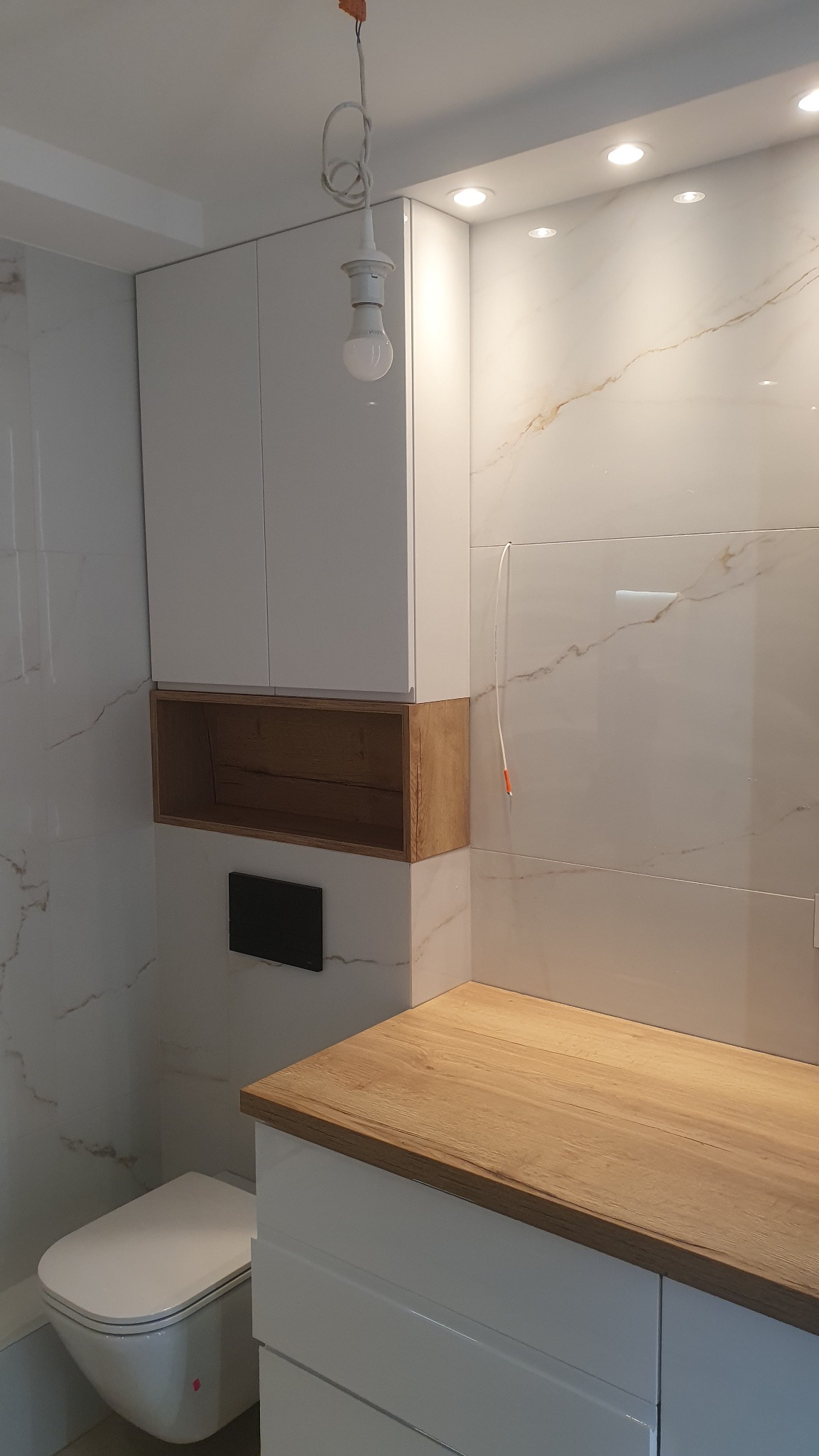 Nowoczesna łazienka z biało-drewnianą zabudową, wisząca toaleta i marmurowe płytki ścienne, widoczne niedokończone podłączenie oświetlenia.