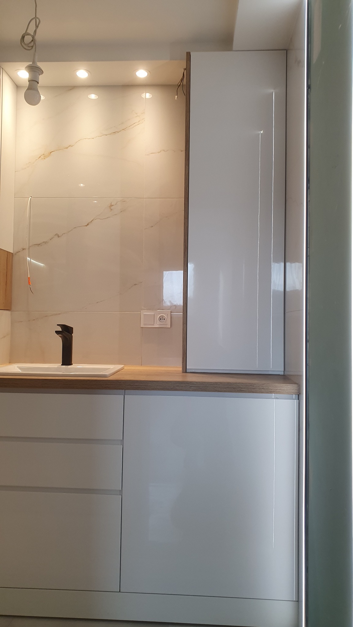 Biała, nowoczesna zabudowa kuchenna z marmurowym blatem i płytkami, czarna bateria umywalkowa, widoczne gniazdko elektryczne, żarówka na kablu, brakujące wykończenie szafki.