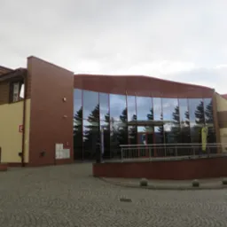 Nowoczesny budynek z dużą, przeszkloną fasadą, w której odbijają się wysokie drzewa iglaste; brukowa droga prowadzi do wejścia.